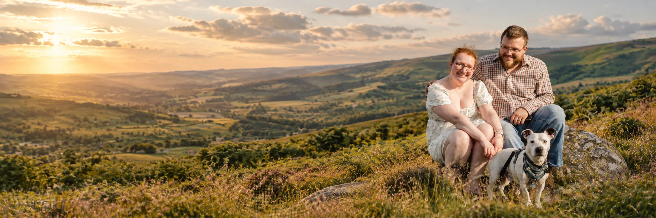 A couple sitting on a hillside at sunset with their dog